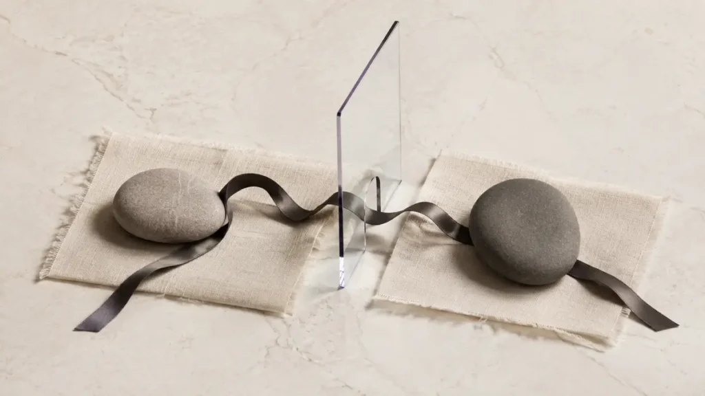 Two stones rest apart on linen, linked by a loose ribbon passing through a clear divider.