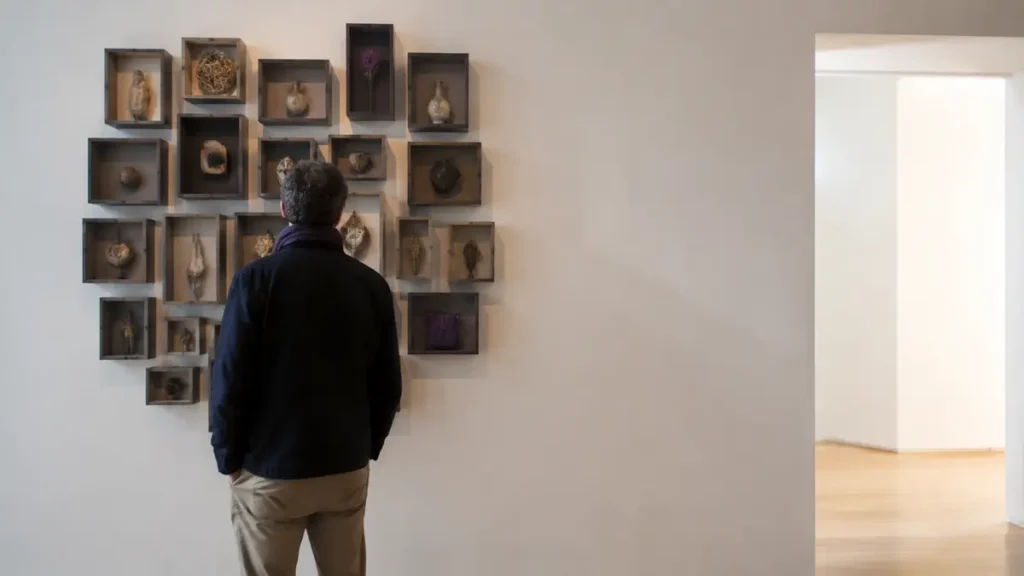 A gallery visitor faces a dense wall of symbolic objects beside a calm, open passage.