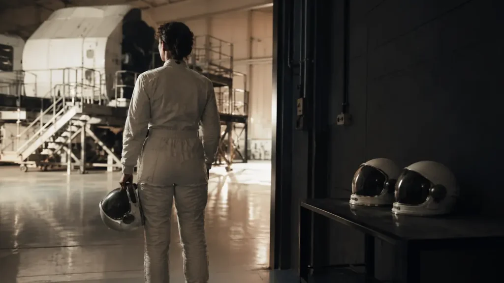 A female astronaut trainee faces a shuttle simulator while two helmets wait together in shadow.
